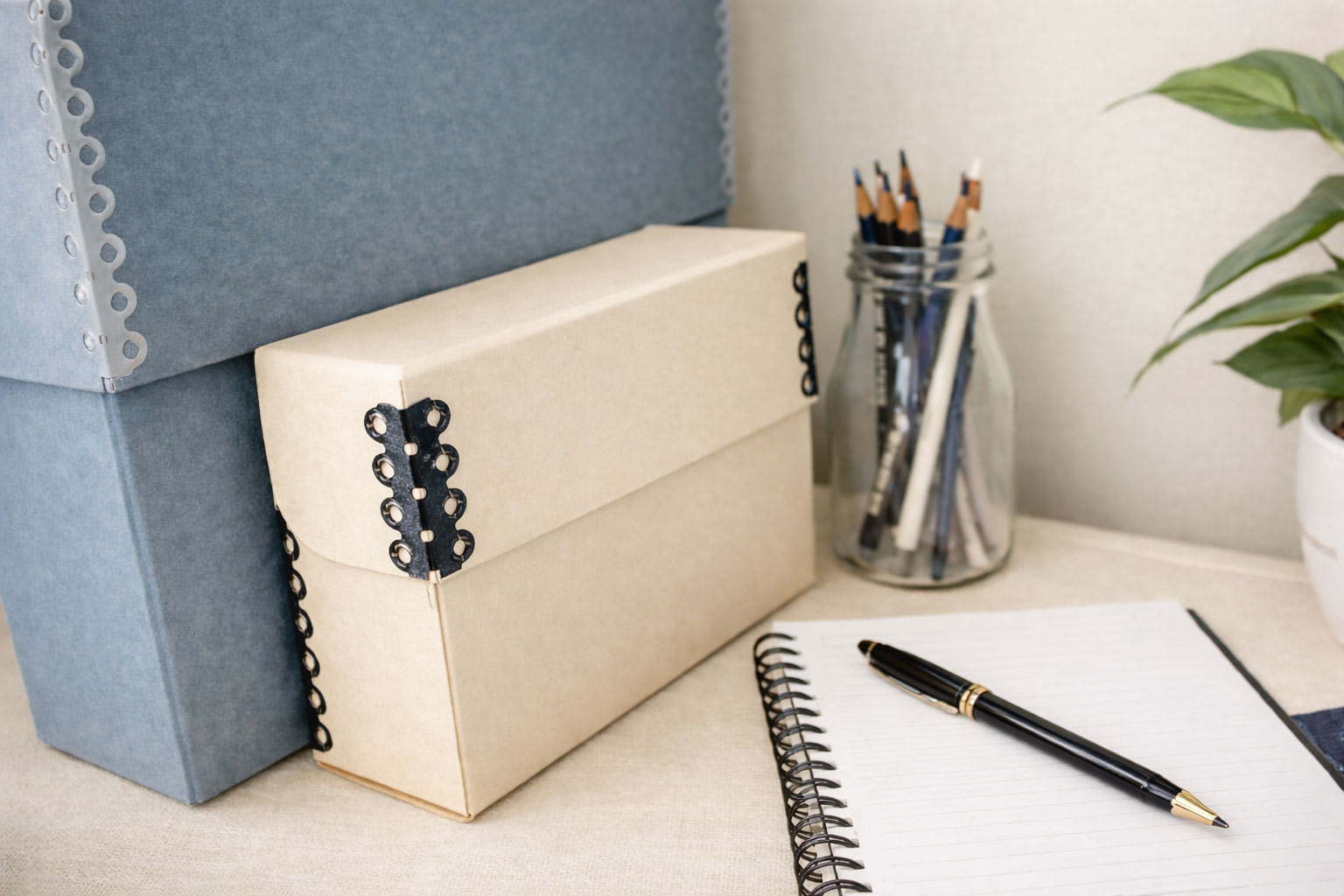 Archival storage boxes beside a notebook, pen, and jar of pencils on a clean workspace, representing thoughtful organization, writing, and care of family archives.